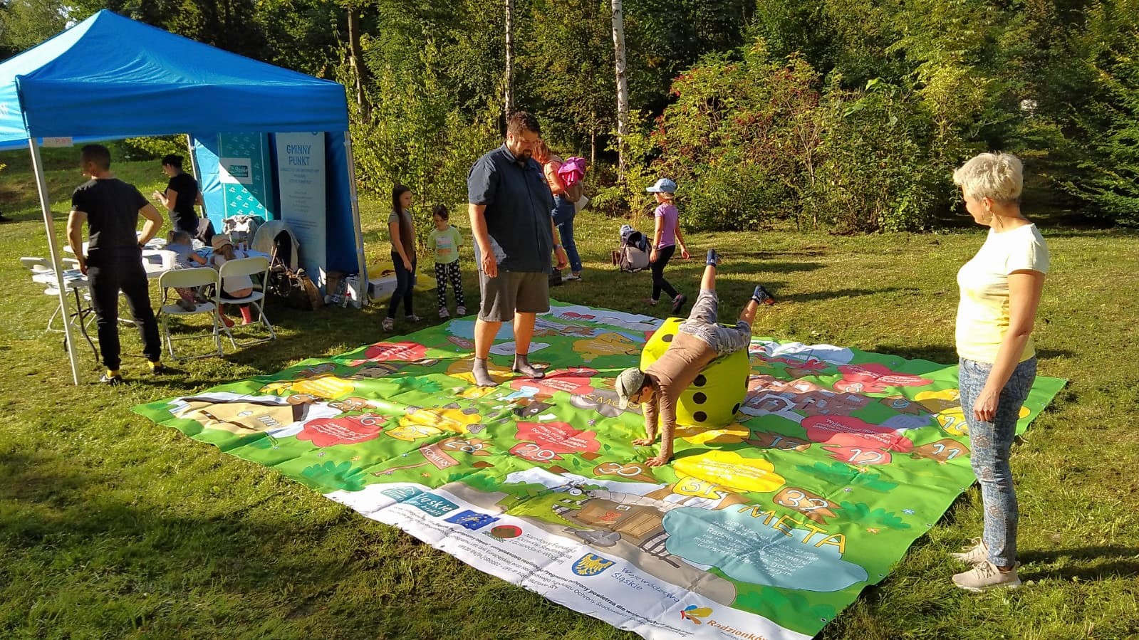 Dwie osoby dorosłe i dwoje dzieci grają w dużą grę planszową rozłożoną na trawie.