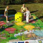 A group of children is playing a large board game laid out on the grass.
