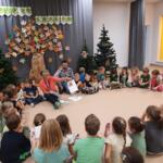 A group of preschoolers sitting with the eco-manager in a circle on the floor inside the preschool classroom. In the background, a decoration mimicking a forest.