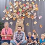 A group of preschoolers sitting with the eco-manager in a circle on the floor inside the preschool classroom. In the background, a decoration mimicking a forest.