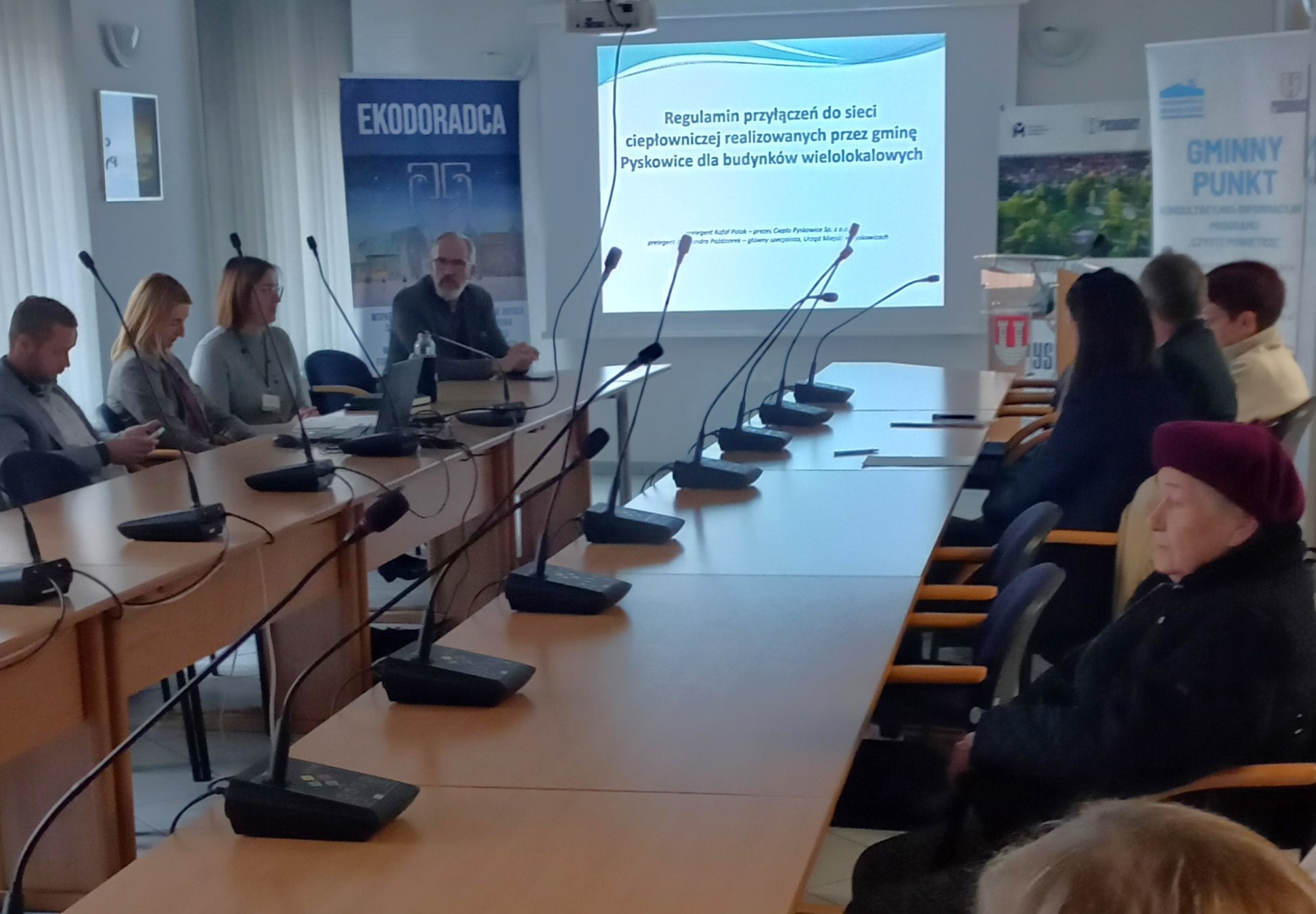 A group of adult participants at the meeting watching presentations displayed on the screen.