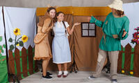 Na zdjęciu scena teatru oraz dwie dziewczynki i mężczyzna na tle dekoracji przedstawiającej dom.